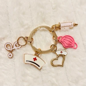 Nyckelring med vårdtema och berlocker - Cool nyckelring i guldmetall med berlocker som stetoskop, spruta, sjuksköterskemössa, hjärta och rosa munskydd. Perfekt accessoar för dig som gillar vård eller pluggar till sjuksköterska.