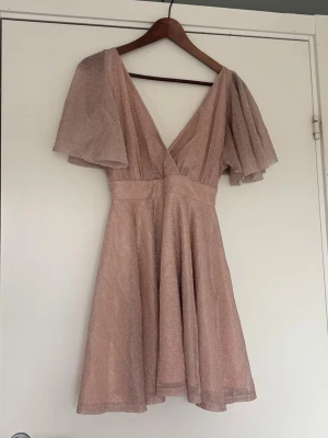 Glittrig kortklänning med v-ringning - Superfin glittrig kortklänning i ljusrosa med v-ringning och vida, korta ärmar. Klänningen har omlott-look upptill och ett markerat midjeband som ger en snygg siluett. 