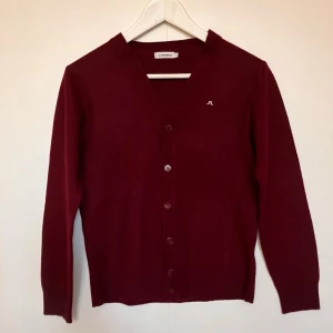 Fin Vinröd cardigan från J.Lindeberg - Fin Vinröd cardigan från J.Lindeberg med knappar framtill och liten broderad logga på bröstet. Klassisk rak modell med långa ärmar och ribbade muddar. Perfekt att slänga över en t-shirt för en snygg lager-på-lager-look. Axelbredd 40cm, längd 58cm, säljes i befintligt skick enligt bilderna!