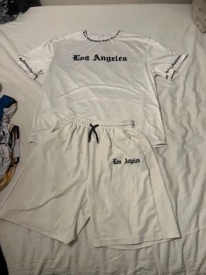 Vitt set med Los Angeles-tryck - Säljer ett vitt matchande set med t-shirt och shorts. T-shirten har svart 'Los Angeles'-tryck framtill och textdetaljer på ärmar och krage. Shortsen har resår i midjan med svart snöre och samma tryck på ena benet. Materialet känns mjukt och sportigt, perfekt för en avslappnad look.