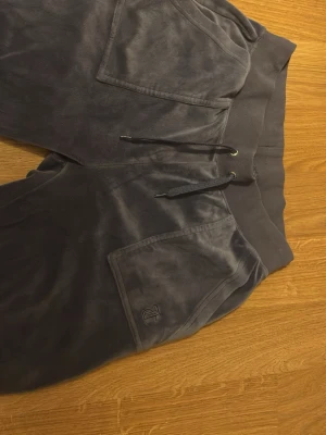 Mörkgrå mjukisbyxor från Juicy Couture - Mörkgrå mjukisbyxor från Juicy Couture i mjuk velour. Byxorna har bred resår i midjan med dragsko och stora fickor framtill, varav ena är broderad med JC-loggan. Perfekta för chill dagar och riktigt skön passform.