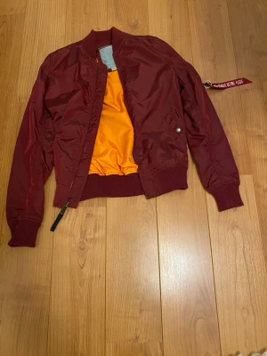 Vinröd bomberjacka från Alpha Industries - Snygg vinröd bomberjacka från Alpha Industries med klassisk orange insida. Jackan har ribbade muddar, dragkedja framtill och en ikonisk röd tag på ärmen. Perfekt för dig som vill ha en streetig och tidlös look.