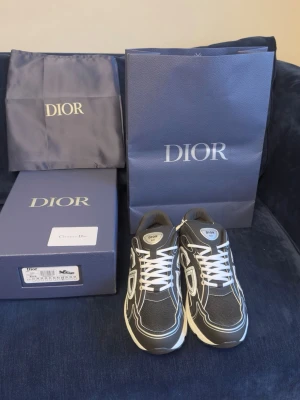 Dior grå sneakers med vita detaljer - Snygga sneakers från Dior i grått mesh och mocka med vita kontrastsömmar och stora CD-loggor på sidorna. Skorna har vita snören, vadderad plös och en chunky vit sula som ger en modern look. Perfekta för dig som vill ha något exklusivt och trendigt.
