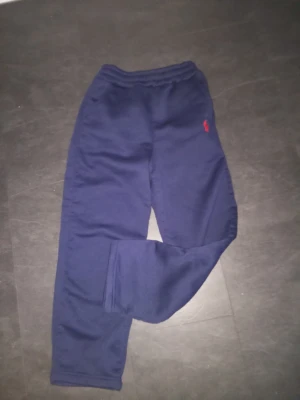 Marinblå mjukisbyxor från Polo Ralph Lauren - Snygga marinblå mjukisbyxor från Polo Ralph Lauren med röd broderad logga på benet. Byxorna har resår i midjan och är gjorda i mjukt bomullsmaterial. Perfekta för en chill och avslappnad stil.