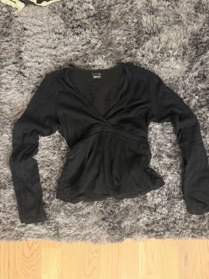 Svart omlottopp från Gina Tricot - Svart omlottopp från Gina Tricot med v-ringning och långa ärmar. Toppen har en figurnära passform och är tillverkad i ett mjukt, ribbat material som ger en snygg struktur. Perfekt att styla med jeans eller kjol. Använt 1 gång!