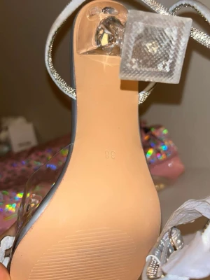 Silvriga pumps med strass och plexiklack - Glittriga pumps i silver med spetsig tå, transparenta detaljer och dekorativa strass på ovandelen. Klacken är i plexiglas med fyrkantig form. Perfekta för dig som vill sticka ut med en trendig och glamorös look.