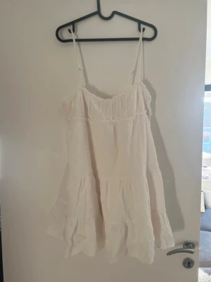 Vit sommarklänning från Zara - Söt vit kortklänning från Zara med tunna axelband och volangdetaljer. Klänningen har en luftig passform och är tillverkad i ett lätt bomullstyg, perfekt för varma dagar. Enkel och stilren design som känns fräsch och trendig.