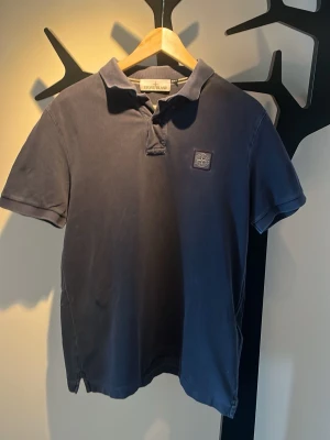 Mörkblå Stone Island piké - Snygg mörkblå piké från Stone Island med klassisk krage och knappslå. T-shirten har korta ärmar och en diskret Stone Island-logga på bröstet. Tillverkad i mjuk bomull för en bekväm känsla och stilren look.