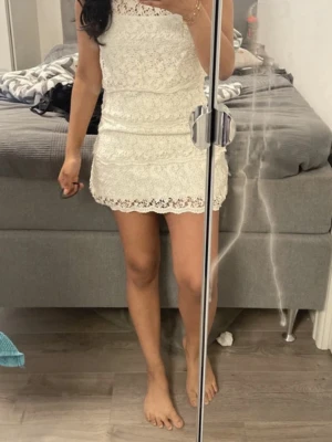 Vit spetsklänning med volang - Söt vit kort klänning i spets med volangdetaljer. Klänningen har en figurnära passform och är perfekt för sommarens tillfällen eller studenten Spetsen ger en romantisk känsla och den korta längden gör den extra trendig.