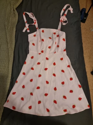 Rosa klänning med jordgubbsmönster - Söt rosa kortklänning med röda jordgubbar och knytbara axelband. Klänningen har en lätt utsvängd form och är perfekt för varma dagar. Materialet känns mjukt och luftigt, och axelbanden ger en lekfull touch.