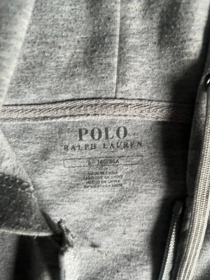 Grå hoodie från Polo Ralph Lauren - Snygg grå hoodie med dragkedja från Polo Ralph Lauren. Klassisk design med huva och dragsnören, två stora fickor framtill och den ikoniska broderade loggan på bröstet. Tillverkad i mjuk bomullsblandning för maximal komfort.
