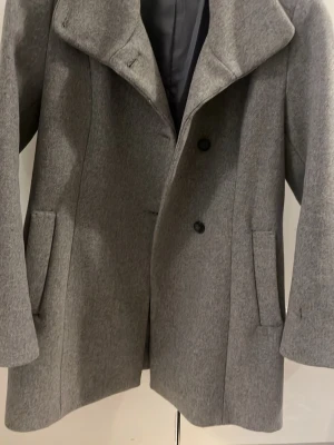 Grå ullkappa med bred krage - Stilren grå kappa i ullblandning med bred krage och två knappar framtill. Kappan har raka ärmar, två djupa fickor och helfodrad insida. Perfekt för kyliga dagar och ger en klassisk look. Den är ifrån Lindex 