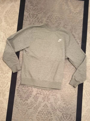 Grå sweatshirt från Nike - Säljer en stilren grå sweatshirt från Nike med klassisk rund hals och diskret Nike-logga på bröstet. Tröjan har ribbade muddar vid ärmslut och nederkant, perfekt för en avslappnad och sportig look.