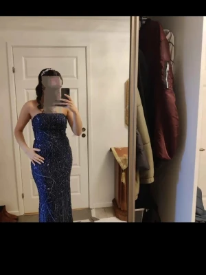Mörkblå glittrig långklänning - Snygg mörkblå långklänning med glittrigt mönster och axelbandslös design. Klänningen har en figurnära passform och är perfekt för speciella tillfällen. Materialet känns lyxigt och skimrar snyggt i ljuset.
