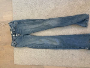 Blå smala jeans med knappar - Snygga blå jeans med bootcut passform och tre synliga knappar i gylfen. Jeansen har klassiska fem fickor och slitningar för en trendig look. Tillverkade i denim med en ljus tvätt och sköna att bära hela dagen.