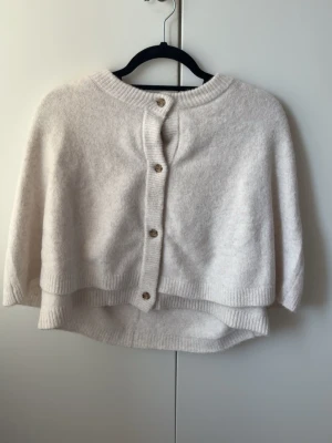 Vit poncho från Kappahl med knappar - Mysig vit poncho från Kappahl med ribbstickade kanter och snygga knappar framtill. Perfekt att slänga över en tröja för extra värme. Mjuk och skön, passar till många olika outfits. Funkar för mig som har Xs/ S, använd en gång 💓