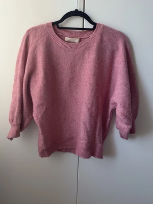 Rosa stickad tröja från Ellos - Mysig rosa stickad tröja från Ellos med rund halsringning och ribbade muddar vid ärmslut och nederkant. Tröjan har en avslappnad passform och något kortare modell med ballongärmar. Perfekt för lager-på-lager och en trendig look.