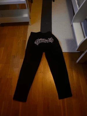 Svarta Hoodrich mjukisbyxor - Svarta mjukisbyxor från Hoodrich med stort vitt och rött logotryck på framsidan. Byxorna har resår och snörning i midjan för skön passform. Perfekta för chill och streetwear-stil.