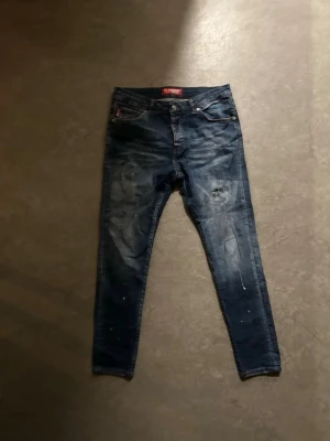 Mörkblåa slimfit dsquared jeans - Säljer ett par mörkblå skinny jeans med slitningar och färgstänk för en cool, avslappnad look. Jeansen har klassisk femficksdesign och låg midja. Perfekta för dig som gillar streetstyle och vill ha ett par jeans med attityd.
