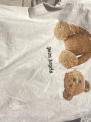 Vit Palm Angels t-shirt med björn - Säljer en vit t-shirt från Palm Angels med ett stort tryck av en nallebjörn på framsidan och Palm Angels-loggan i svart på både fram- och baksidan. Klassisk passform och tillverkad i mjuk bomull. Perfekt för dig som gillar streetwear och coola prints.