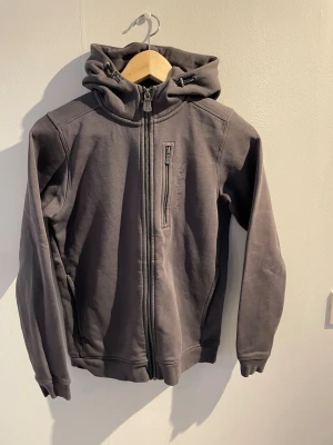 Mörkgrå hoodie med dragkedja - Snygg mörkgrå hoodie från Peak Performance med hel dragkedja framtill och en bröstficka med dragkedja. Tillverkad i mjukt material och har en stor huva med broderad logga bak. Perfekt för en avslappnad och sportig stil.