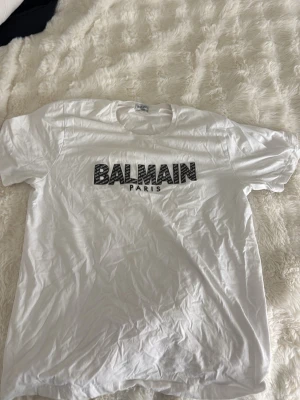 Vit Balmain Paris t-shirt - Säljer en vit t-shirt från Balmain Paris med svart logotyptryck på bröstet. Klassisk passform med rund hals och korta ärmar. Perfekt för dig som gillar stilrena och trendiga plagg med exklusiv känsla.