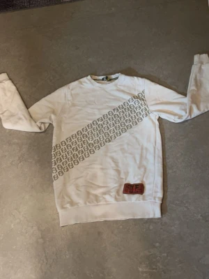 Vit Fendi hoodie med logomönster - Säljer en vit hoodie från Fendi med diagonal logomönster över bröstet och en röd patch längst ner. Tröjan har rund halsringning och ribbade muddar. Perfekt för dig som gillar exklusiva märken och stilren design.