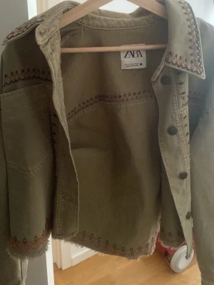 Olivgrön jeansjacka från Zara - Snygg olivgrön jeansjacka från Zara med dekorativa broderier i rött och svart längs krage, ärmslut och nederkant. Jackan har råa kanter och en croppad passform som ger en trendig look. Perfekt för dig som vill sticka ut lite extra.