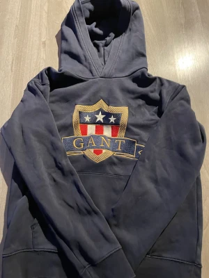 Mörkblå hoodie från GANT med emblem - Snygg mörkblå hoodie från GANT med stort broderat emblem på bröstet i guld, rött, vitt och blått. Klassisk huva och känguruficka framtill. Perfekt för dig som gillar preppy stil och vill ha något bekvämt men ändå stilrent.