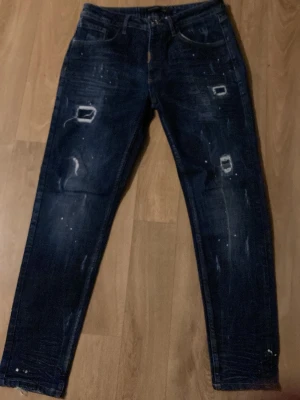 Mörkblå alessandro zavetti jeans - Säljer ett par mörkblå jeans med slitna detaljer och vita färgstänk. Jeansen har raka ben, klassisk femficksmodell och coola revor framtill för en trendig look. Perfekta för dig som gillar streetstyle och vill sticka ut lite extra.