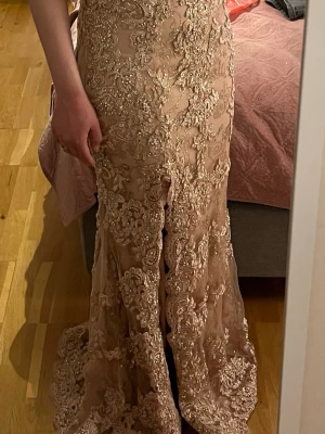 Champagnefärgad långklänning med spets - Elegant långklänning i champagnefärg med vackra spetsdetaljer och broderade mönster. Klänningen har figurnära passform och en slits framtill som ger en extra touch. Perfekt för dig som vill sticka ut på bal eller fest.