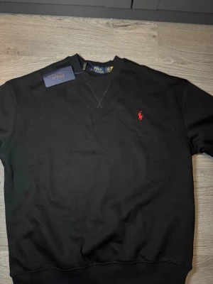 Svart sweatshirt från Polo Ralph Lauren - Säljer en svart sweatshirt från Polo Ralph Lauren med klassisk rund hals och ribbade muddar. Tröjan har det ikoniska röda Polo-logot broderat på bröstet och är tillverkad i mjukt bomullsmaterial. Perfekt för en avslappnad och stilren look.