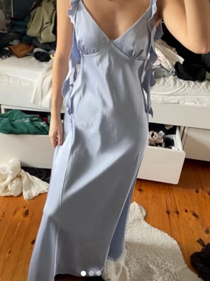 Ljusblå långklänning med volanger - Långklänning i ljusblå färg med v-ringad front och smala axelband. Klänningen har dekorativa volanger längs sidorna och är ärmlös. Materialet ser ut att vara bomull eller polyester, vilket ger en mjuk och lätt känsla.