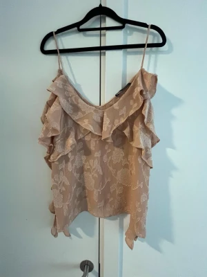 Beige blommig linnetopp med volang från Zara  - Söt beige linnetopp med tunna axelband och volangdetaljer längs ringningen och sidorna. Toppen har ett blommigt mönster i ljusa toner och är tillverkad i ett lätt, luftigt material som passar perfekt till sommaren.