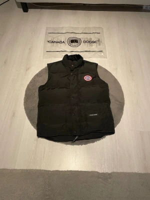  Canada Goose väst svart  - Säljer en svart, vadderad väst från Canada Goose med hög krage och tryckknappar framtill. Västen har det klassiska Canada Goose-märket på bröstet och innerfickor för extra förvaring. Perfekt för lager-på-lager under kyliga dagar.