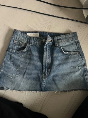 Blå jeanskjol från Zara - Säljer en klassisk blå jeanskjol från Zara med rå kant nertill och slitna detaljer. Kjolen har fem fickor, bälteshällor och stängs med knapp och dragkedja. Perfekt till en avslappnad look.