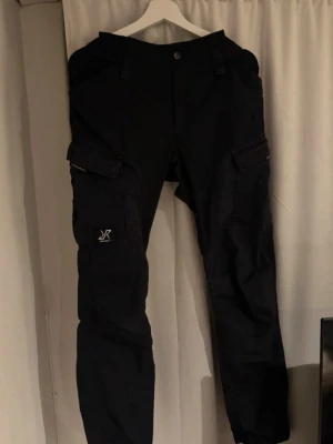 Svarta cargobyxor RevolutionRace - Svarta cargobyxor från RevolutionRace med flera praktiska fickor på benen och klassisk cargomodell. Byxorna har en rak passform och är tillverkade i slitstarkt material, perfekta för en aktiv livsstil. Snygg logga på ena benfickan.