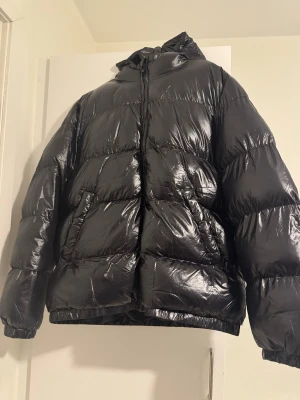 Svart glansig pufferjacka med huva - Säljer en svart pufferjacka med glansig finish och huva. Jackan har breda kanaler, dragkedja framtill och elastiska muddar vid ärmslut och nederkant. Perfekt för kalla vinterdagar och riktigt trendig look.