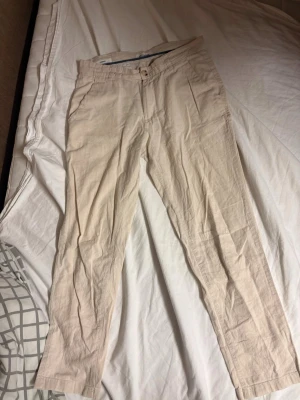 Beiga linnebyxor från Jack & Jones - Snygga beiga linnebyxor med rak passform och klassisk design. Byxorna har fickor i sidorna och stängs med knapp och dragkedja. Perfekta för en avslappnad och stilren look. Tillverkade i ett lätt och luftigt linnematerial. Och som ni ser på sista bilden har det skett en reparation