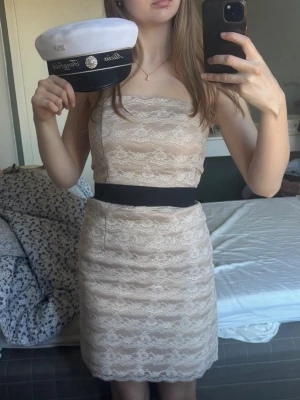 Vit spetsklänning - studentklänning- bandeuklänning  - Söt axelbandslös kortklänning i beige med vit spets och svart band i midjan. Klänningen har en figurnära passform och bandeutopp. 