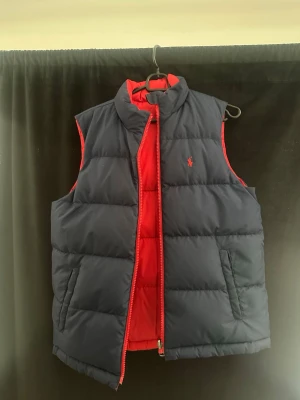 Vändbar dunväst från Polo Ralph Lauren - Snygg vändbar dunväst från Polo Ralph Lauren i marinblått och rött. Västen har hög krage, tvåvägsdragkedja och två fickor på varje sida. Klassisk broderad logga på bröstet. Perfekt för lager-på-lager och stilren look.