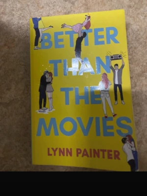 Better Than the Movies bok - En färgglad pocketbok med titeln 'Better Than the Movies' av Lynn Painter. Omslaget är gult med blå text och illustrationer av romantiska par och en person med boombox. Perfekt för dig som gillar romantisk ungdomslitteratur.