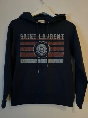 Mörkblå Saint Laurent hoodie - Snygg mörkblå hoodie från Saint Laurent med tryck framtill i vitt och rött, samt stor känguruficka och justerbar huva. Perfekt för en avslappnad och trendig stil. Materialet känns mjukt och bekvämt mot huden.