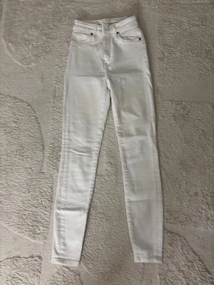 Vita skinny jeans med hög midja - Säljer ett par vita skinny jeans med hög midja och klassisk femficksdesign. Jeansen har smal passform och stängs med dragkedja och knapp. Perfekta för en clean och fräsch look.