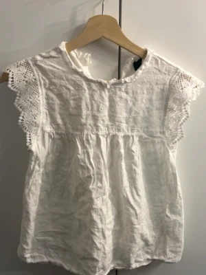 Vit blus med spetsdetaljer - Söt vit blus med korta ärmar och vackra spetsdetaljer längs ärmsluten. Blusen har en luftig och lätt struktur med rynk under bysten för en avslappnad passform. Den måste dock strykas 