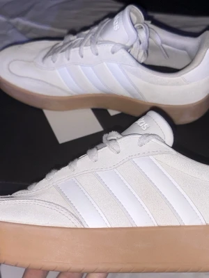 Vita Adidas sneakers med gummisula - Fräscha vita sneakers från Adidas med klassiska tre ränder på sidan och snygg ljusbrun gummisula. Ovandelen är i mockaliknande material och detaljer i läder. Perfekta för en clean och stilren look. Har använts bara få gånger 