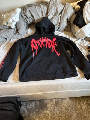 Svart Revenge hoodie med röd print - Säljer en svart hoodie från Revenge med stor röd logga i gotisk stil på bröstet och en stor röd handavtrycksdesign på ryggen. Hoodien har huva och känguruficka framtill samt tryck på ena ärmen. Perfekt för dig som gillar streetwear och statement-plagg.