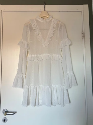 Vit volangklänning - studentklänning  - Supersöt vit kortklänning från Missguided med volanger och hög krage. Klänningen har långärmade ärmar med volangdetaljer och är tillverkad i ett prickigt mesh tyg 