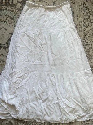 Vit långkjol - Säljer en vit långkjol i mjukt material med volangdetaljer. Kjolen har en enkel, flowy passform och resår i midjan för extra komfort. Perfekt för dig som gillar en avslappnad och stilren look. Lite nopprig på insidan men det syns inte utåt