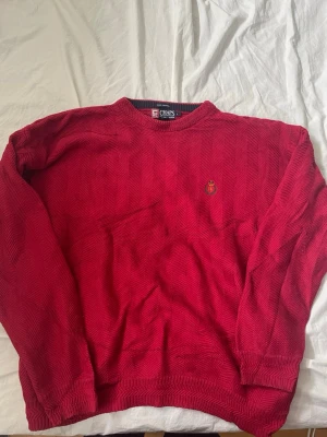 Röd stickad tröja från Ralph Lauren x Chaps - Snygg röd stickad tröja från Chaps x Rakph Lauren med diskret fiskbensmönster och broderad logga på bröstet. Tröjan har rund halsringning och ribbade muddar vid ärmslut och nederkant. Perfekt för dig som gillar klassisk stil.